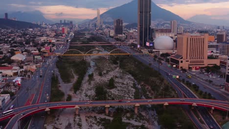 Drone-ascending-over-the-Santa-Catarina-river-at-sunset-reveals-busy-downtown-area-of-Monterrey,-Mexico-with-bright-sign-boards-and-bridges-crossing-the-dry-riverbed
