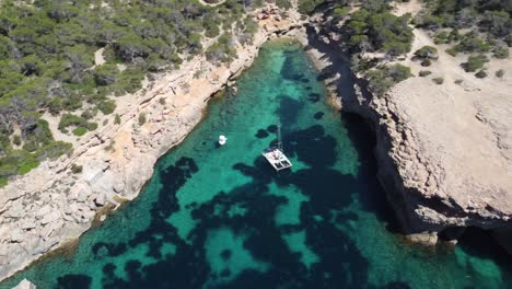 Drone-aerial-shot-view-of-a-catamaran-peacefully-anchored-in-a-turquoise-cove-of-Ibiza,-basking-in-the-sunny-bliss-of-a-perfect-day-at-sea