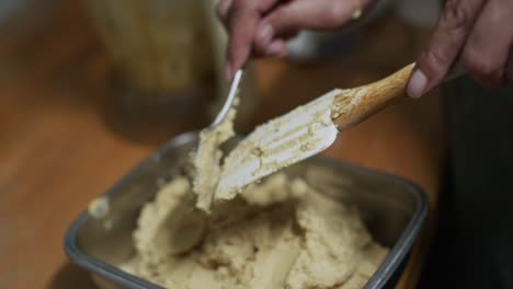 Freshly-made-hummus-paste-scraped-off-baking-spoon-into-metal-container,-filmed-as-close-up-slow-motion-shot