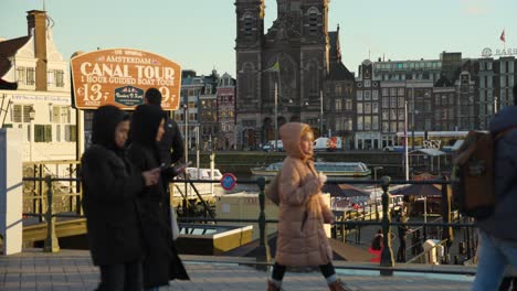 Tourists-Walking-past-Canal-Boat-Tour-Waterfront-In-Amsterdam-On-Cold-Autumnal-Day