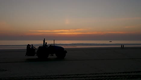 Tractor-passing-on-the-beach-at-sunset-with-people-in-the-distance,-seagulls,-a-fishing-boat-in-the-distance-and-surfers-in-the-water