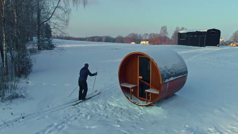 Person-on-cross-country-skies-skiing-past-a-sauna-and-to-a-trailer-cabin---aerial-follow