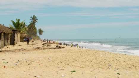 People-work-on-sand-beach-by-houses-in-Moree-village-in-Ghana,-wide