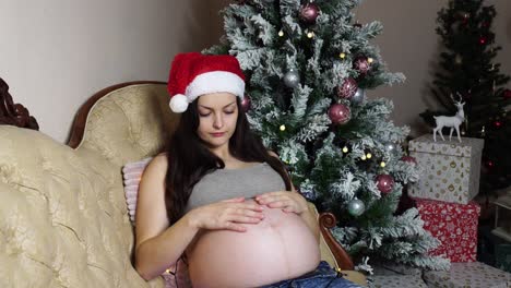 Schwangere-Mädchen-Tragen-Weihnachtsmütze-Und-Berühren-Runden-Nackten-Bauch-In-Der-Nähe-Des-Weihnachtsbaums