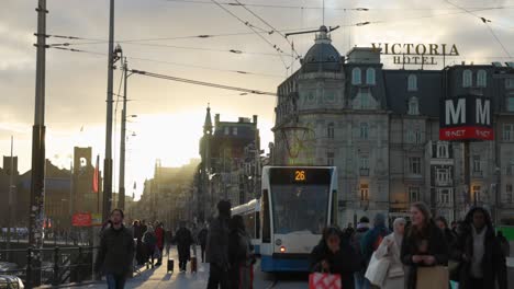 beautiful-Amsterdam-building-in-the-distance,-as-a-blue-and-white-public-tram-passes-by-transporting-commuters-around-the-busy-city,-Netherlands-during-Golden-hour