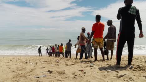 Men-pull-up-fishing-net-by-rope-to-sand-beach-in-Ghana,-from-behind