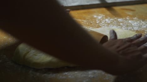 Freshly-raised-dough-pressed-flat-using-rolling-pin-on-wooden-kitchen-top-by-chef,-filmed-as-close-up-slow-motion-shot