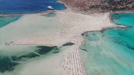 Drone-view-in-Greece-flying-over-balos-beach-with-clear-blue-sea-on-the-sides-and-white-sand-surrounded-by-brown-landscape-on-a-sunny-day-in-Crete