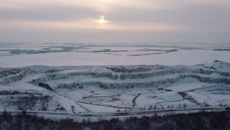 Aerial-view-of-a-snow-covered-quarry-pit-in-winter-evening-with-sun-peeking-out