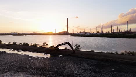 Silhouette-of-excavator-next-to-large-lake-with-oil-refinery-stacks-backlit-by-sunset-on-island