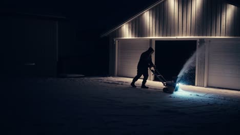 Man-Clearing-Snow-Outside-Cabin-With-Snow-Blower-At-Night