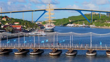 Blue-flags-from-marathon-wave-in-wind-on-Queen-Emma-bridge-as-large-masted-ship-in-port-is-in-front-of-Queen-Juliana-Bridge