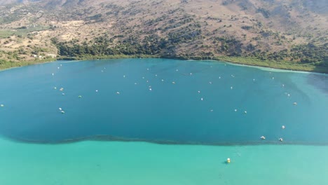 Vista-De-Drones-En-Grecia-Volando-Sobre-Un-Lago-Azul-Claro-Y-Oscuro-Con-Pequeñas-Embarcaciones-Y-Rodeado-De-Montañas-Verdes-En-Un-Día-Soleado-En-Creta