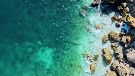 Clear-ocean-water-shimmers-as-it-crashes-on-rocky-coast,-nature-background-drone-top-down