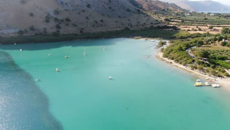 Drone-view-in-Greece-flying-over-a-light-and-dark-blue-lake-with-small-boats-and-surrounded-by-green-mountain-on-a-sunny-day-in-Crete