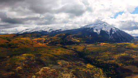 Dramatic-sunny-cloudy-autumn-Aspen-Tree-fall-colors-Kebler-Pass-aerial-cinematic-drone-snow-on-peaks-landscape-Crested-Butte-Gunnison-Colorado-early-fall-red-yellow-orange-Rocky-Mountains-reveal-back