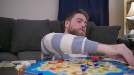 A-bearded-man-plays-the-board-game-Catan-in-his-living-room,-rolling-dice-then-drawing-resource-cards-and-adding-them-to-his-hand