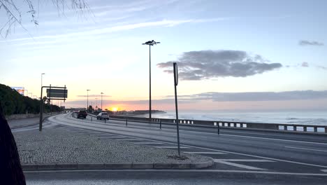 Fantastischer-Sonnenaufgang-Hinter-Der-Festung-In-Carcavelos,-Mit-Autos,-Die-Entlang-Der-Uferpromenade-Fahren