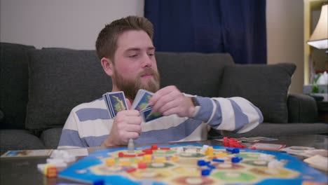 A-bearded-man-plays-the-board-game-Catan-in-his-living-room,-laying-cards-down-then-playing-pieces-on-the-board-and-smiling-and-laughing