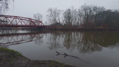 A-small,-picturesque-arched-red-steel-bridge,-as-seen-from-a-little-park,-crossing-a-small-river-in-upstate-New-York-on-a-rainy,-dreary,-early-spring-day