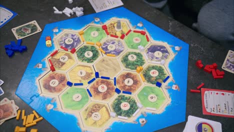 45-degree-overhead-view-of-Catan-gameplay-with-four-players-on-stone-table-with-dice-rolling,-road-building,-and-settlement-placing-by-blue-player-hand