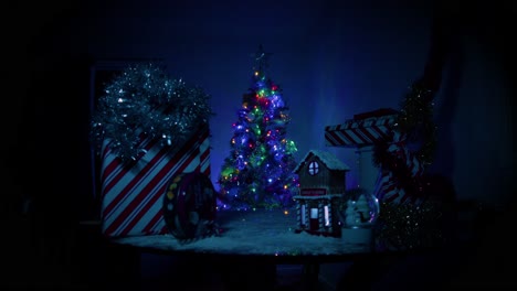 Extreme-Weitwinkelaufnahme-Der-Kamera,-Die-Sich-Auf-Eine-Kleine-Anordnung-Eines-Niedlichen-Weihnachtsdorfes-Im-Schnee-Zubewegt,-Während-Ein-Großer,-Schöner,-Gut-Beleuchteter-Weihnachtsbaum-Im-Fokus-Hinter-Der-Mini-Dorfanordnung-Steht
