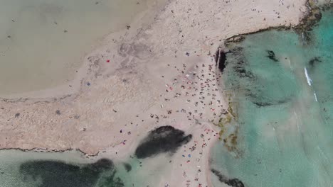 Drone-view-in-Greece-top-view-over-balos-beach-with-clear-blue-sea-on-the-sides-and-white-sand-with-people-swimming-on-a-sunny-day-in-Crete