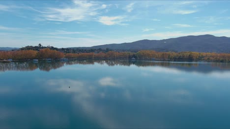 Vuelo-De-Drones-Sobre-El-Lago-De-Banyoles-Y-El-Agua-Azul-Que-Refleja-El-Cielo-En-Otoño
