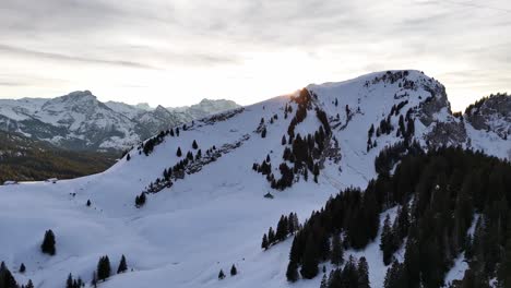 Aerial-drone-shot-revealing-sunlight-of-sunrise-behind-snowy-Gulmen-Mountain-in-Switzerland