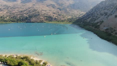 Drone-view-in-Greece-flying-over-a-light-and-dark-blue-lake-with-small-boats-and-surrounded-by-green-mountain-on-a-sunny-day-in-Crete