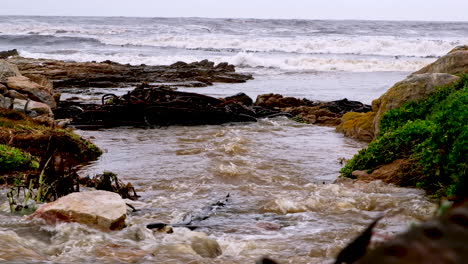 Murky-rainwater-runs-into-rough-sea-on-coastline-from-drainage-system,-telephoto