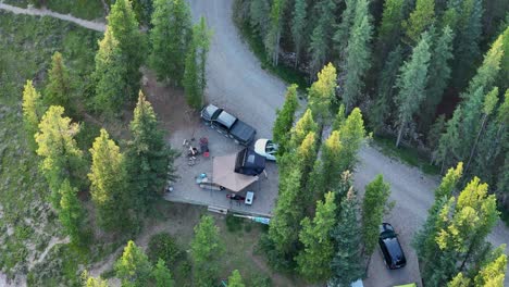 Birds-eye-view-of-a-campsite-with-a-rooftop-tent-setup-and-a-couple-vehicles-surrounded-by-large-pine-trees