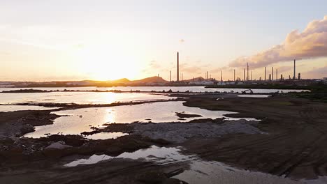 Aerial-dolly-above-asphalt-lake-and-white-piles-of-refuse-at-sunset-with-oil-refinery-stacks-in-distance