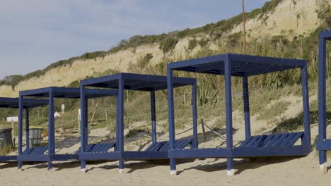 Leere-Blaue-Strandzelte-Aus-Holz-Mit-Einer-Klippe-In-Der-Ferne-Am-Strand-Von-Fonte-Da-Telha-In-Portugal
