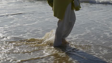 Woman-with-a-green-sweater-tied-around-her-waist-and-white-pants-walks-along-the-seashore-dragging-her-bare-feet