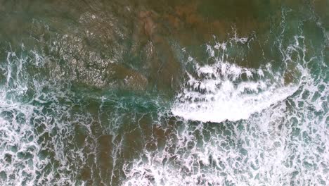 Aerial-view-of-a-wave-breaking-on-a-golden-sandy-beach,-The-mesmerizing-patterns-of-foam-and-surf