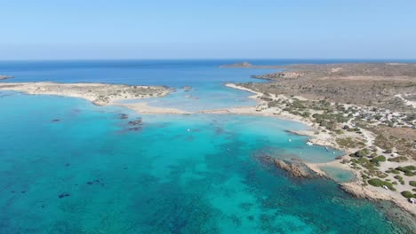 Drone-view-in-Greece-flying-over-Elafonisi-white-sand-narrow-beach,-clear-blue-water-on-the-sides-and-brown-landscape-on-a-sunny-day