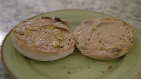 Drizzle-and-spread-honey-on-a-toasted-English-muffin-with-peanut-butter-on-a-green-plate-on-a-marble-countertop