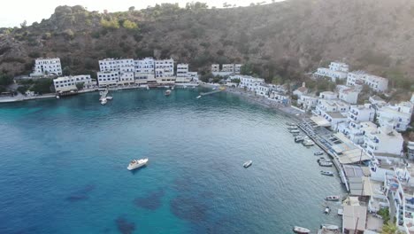 Drone-view-in-Greece-panning-up-over-clear-blue-sea-in-Loutro-small-white-house-town-and-small-boats-next-to-a-hill-on-a-sunny-day