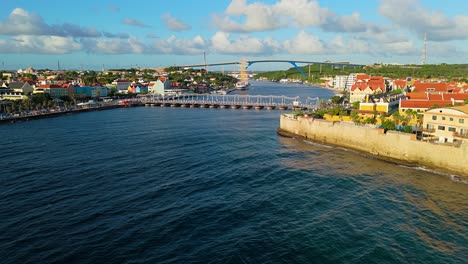 Willemstad-Curacao-Al-Atardecer-Mientras-El-Agua-Del-Océano-Brilla-Con-Un-Velero-Con-Mástiles-En-El-Puerto