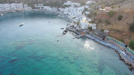 Drone-view-in-Greece-flying-over-clear-blue-sea-in-Loutro-small-white-house-town-and-small-boats-next-to-a-hill-on-a-sunny-day