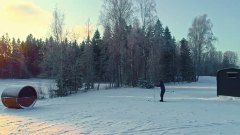 Cross-country-skiing-from-a-countryside-trailer-cabin-and-a-sauna---low-altitude-aerial-follow-at-sunrise