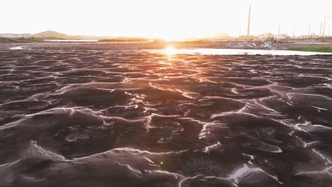 Sunlight-glistens-against-polluted-water-in-asphalt-lake-at-golden-hour-with-oil-refinery-behind-barren-ground