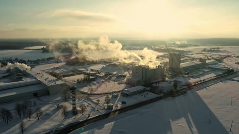 Backlit-smoke-rising-from-industrial-building-covered-in-snow-backlit-by-sun-in-winter