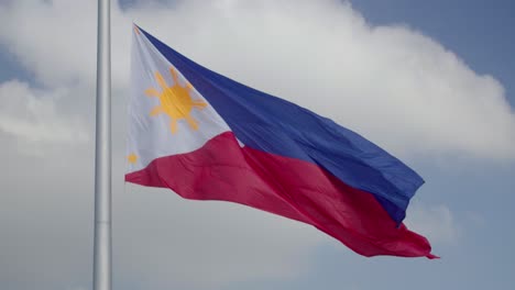 Gran-Bandera-Filipina-Ondeando-En-El-Viento-Contra-Un-Cielo-Parcialmente-Nublado-Mientras-Está-En-Un-Gran-Asta-De-Bandera-En-Cámara-Lenta-De-4k