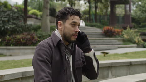Push-in-on-handsome-Hispanic-ethnicity-man-talks-positively,-confident-on-the-phone-outdoors-in-the-park