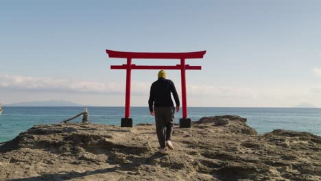 Young-male-walking-toward-Japanese-red-torii-gate-in-front-of-ocean