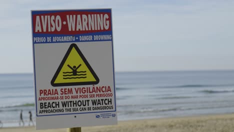 Warning-of-danger-of-drowning-in-portuguese-and-english-on-Fonte-da-Telha-beach-in-Portugal-with-cyclist,-couple-and-dog-in-the-distance