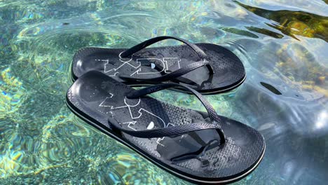 Floating-black-Lidl-supermarket-flip-flops-in-transparent-turquoise-sea-water-by-the-beach-in-Spain,-fun-sunny-summer-vacation-holiday-shoes,-4K-shot