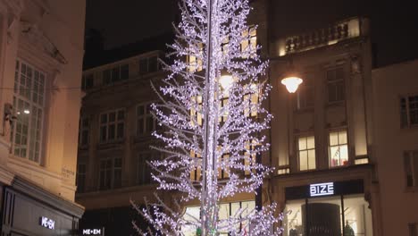 Nach-Unten-Geneigte-Aufnahme-Eines-Beleuchteten-Festlichen-Weihnachtsbaums-Entlang-Einer-Straße,-An-Der-Nachts-Passanten-Vorbeikommen